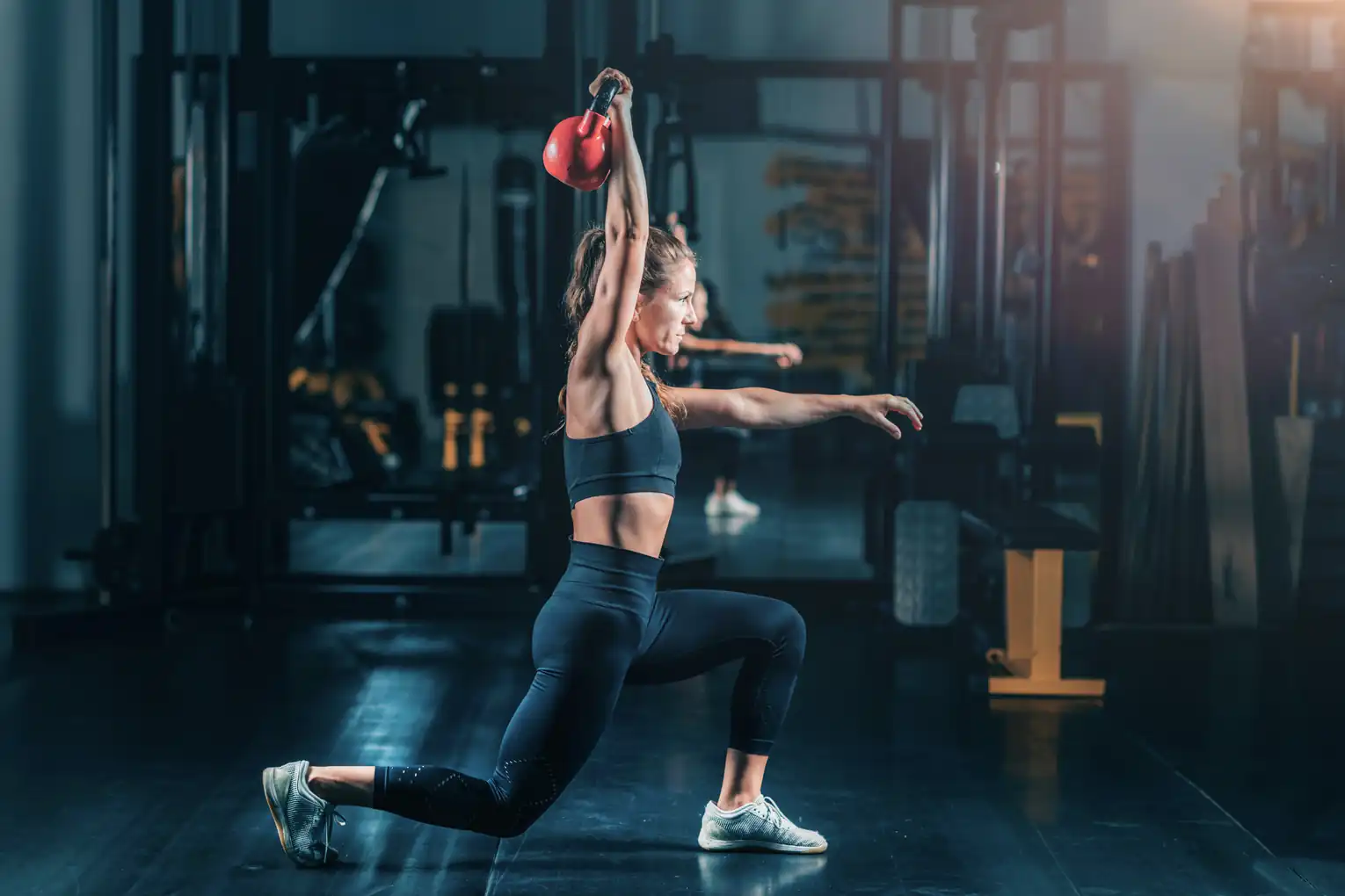 Woman performing a red kettlebell overhead lunge, representing functional strength training, cross-training routines, mobility exercises, and personal health coaching.