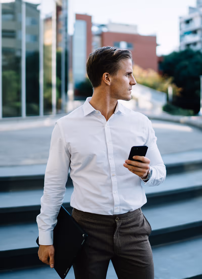Ein Mann in einem weißen Hemd, der ein Smartphone und eine schwarze Mappe in der Hand hält und draußen in der Nähe einer Treppe und moderner Gebäude steht.