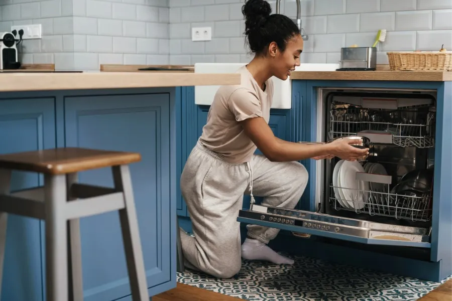 Woman kneeling in a blue kitchen while loading a dishwasher with dishes.