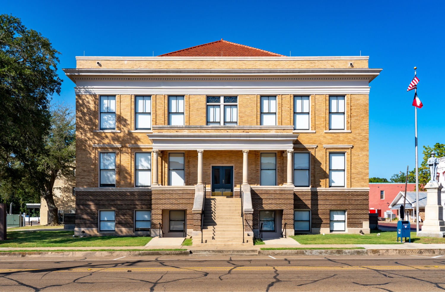 Three-story historic brick courthouse with front steps, columns, and flags under clear blue sky.