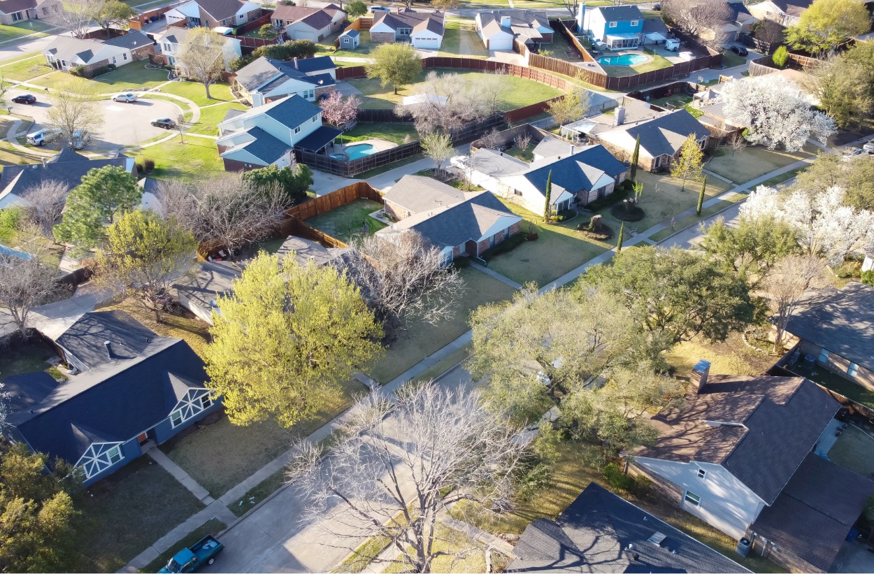 Aerial view of a suburban neighborhood with houses, trees, fenced backyards, and some pools in spring.