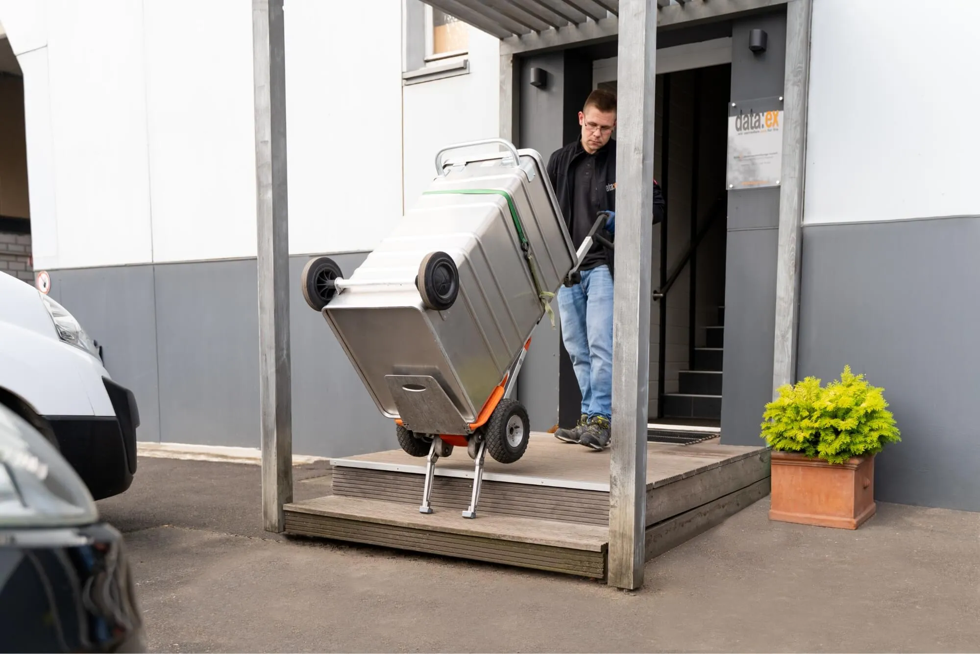 Foto eines Mitarbeiters der Firma data:ex der einen Datenschutzbehälter mittels Sackkarre aus einem Gebäudeeingang transportiert.