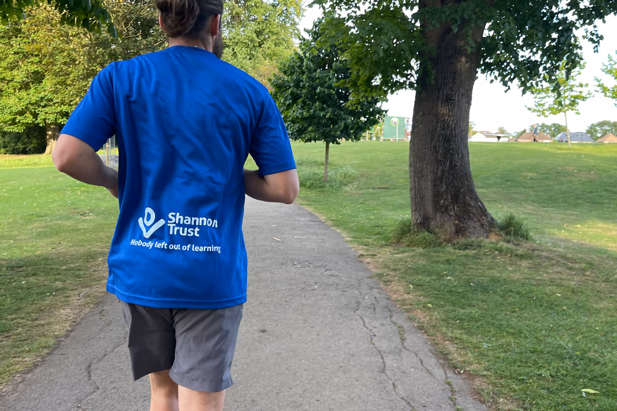 Person jogging on a park path wearing a blue shirt with 'Shannon Trust Nobody left out of learning' printed on the back.