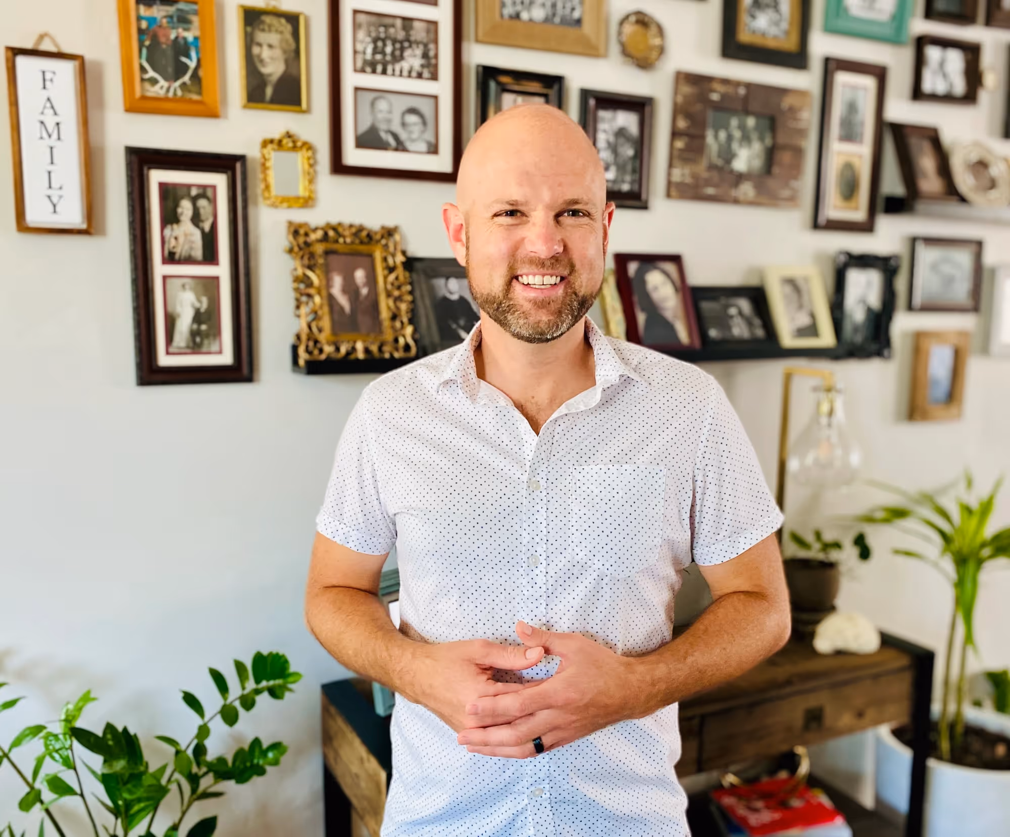 Joseph B. Shumway standing indoors in front of a wall of framed family photos, with plants and a wooden console table in the background.