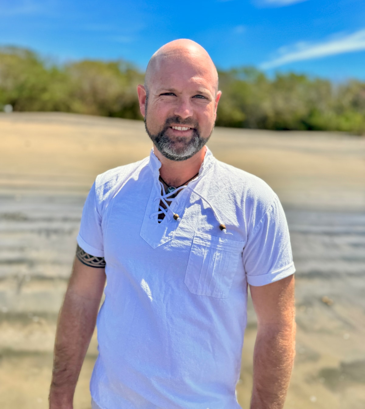 Joseph in white shirt standing on a beach with trees and blue sky background.