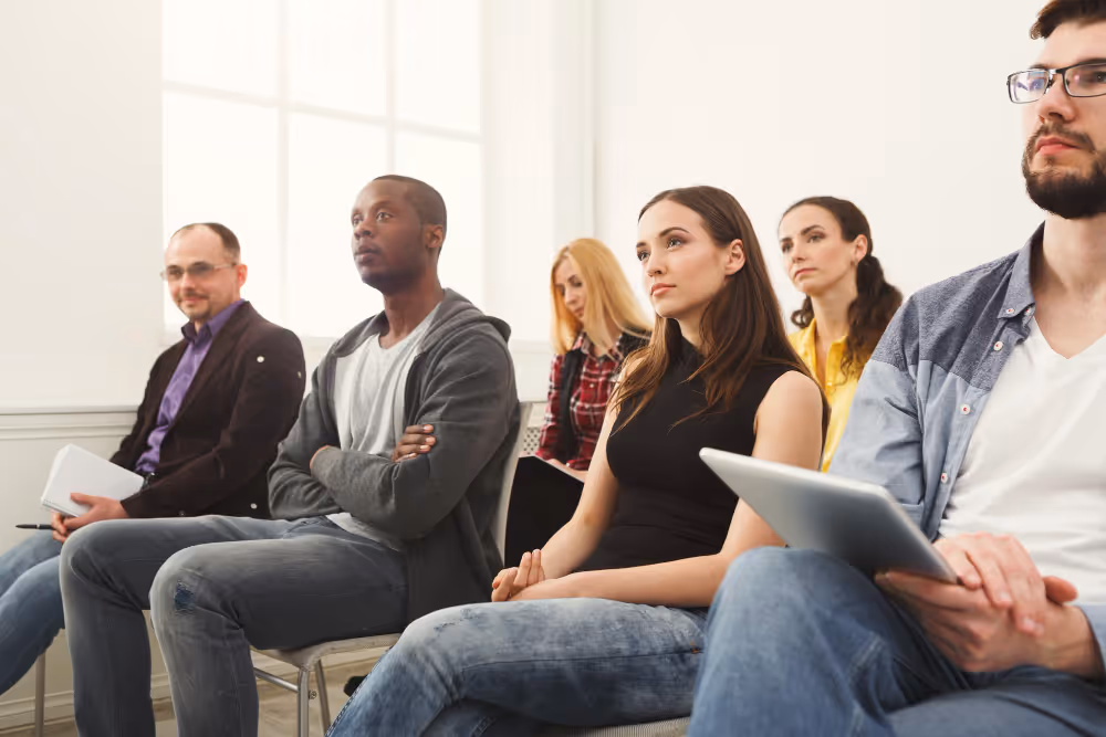 Group of people seated in a bright room, listening attentively during a presentation; one person holds a tablet.