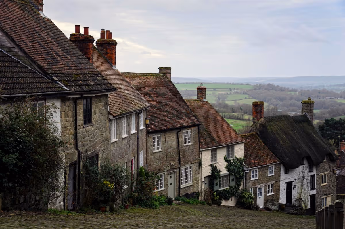 Stone cottages with steep tiled and thatched roofs on a hillside, overlooking green countryside under a cloudy sky.