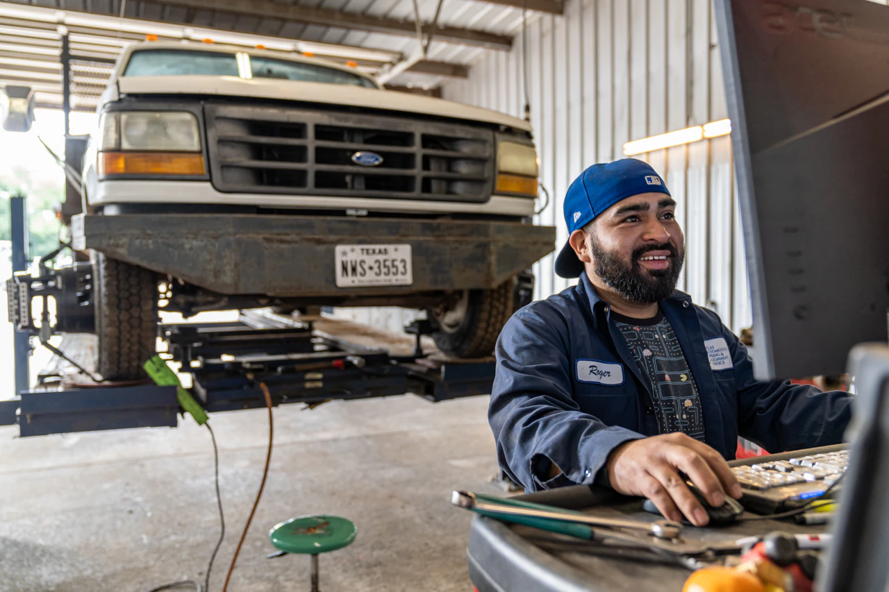 A smiling mechanic wearing a blue cap and work uniform operates a computer inside a truck repair shop. Behind him, a white Ford pickup is lifted on a hydraulic platform for service, symbolizing professional diagnostics and maintenance efficiency in action.