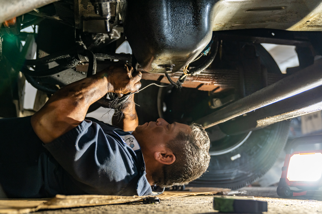 Mechanic performing undercarriage repairs on a heavy-duty truck using hand tools and portable lighting in a repair shop.