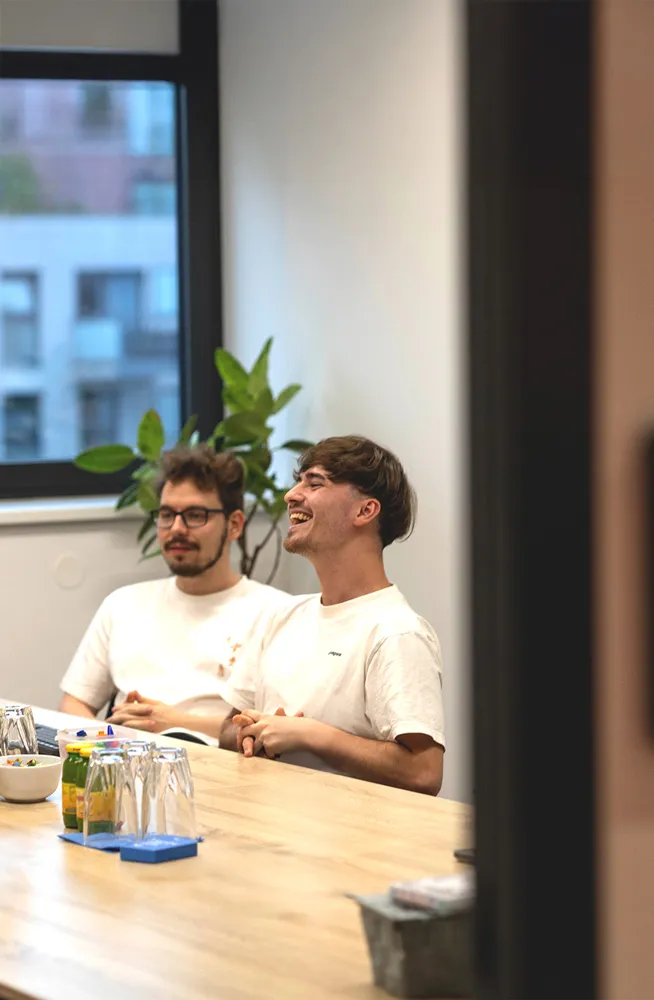 Zwei junge Männer sitzen an einem Besprechungstisch, einer lacht, während der andere aufmerksam ist.