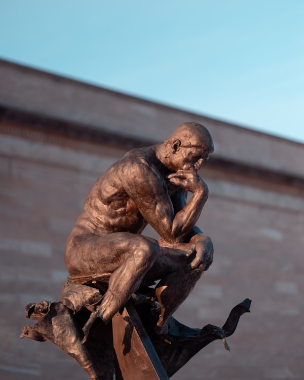 A dramatic close-up of a bronze sculpture reminiscent of Rodin's "The Thinker," perched atop twisted metal with a contemplative pose against a soft blue sky and stone building backdrop.