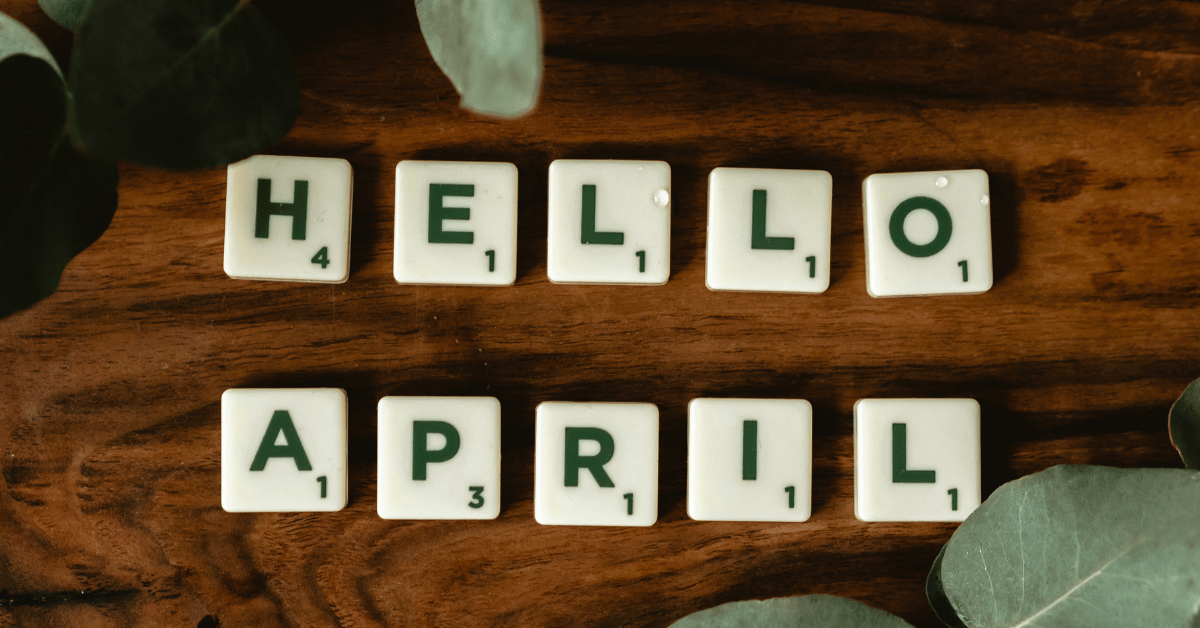 Scrabble tiles spelling "HELLO APRIL" on a wooden surface, surrounded by green leaves.