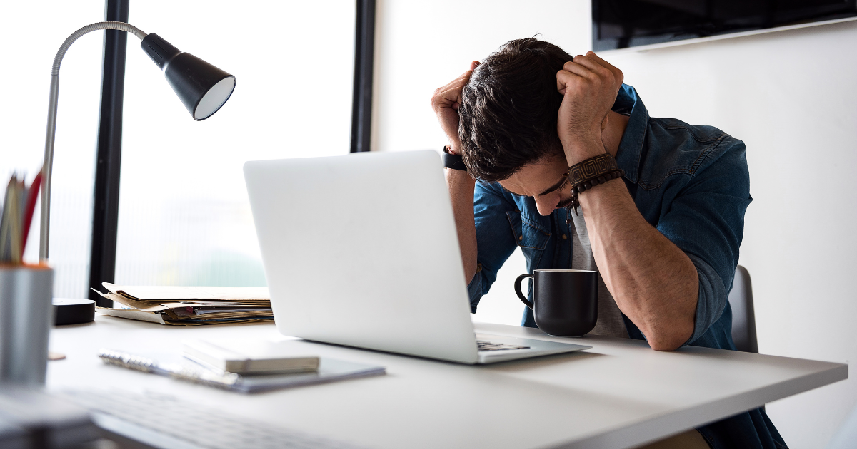 A person sitting at a desk with their head in their hands, appearing stressed or upset. A laptop, a lamp, and a stack of papers are on the desk.