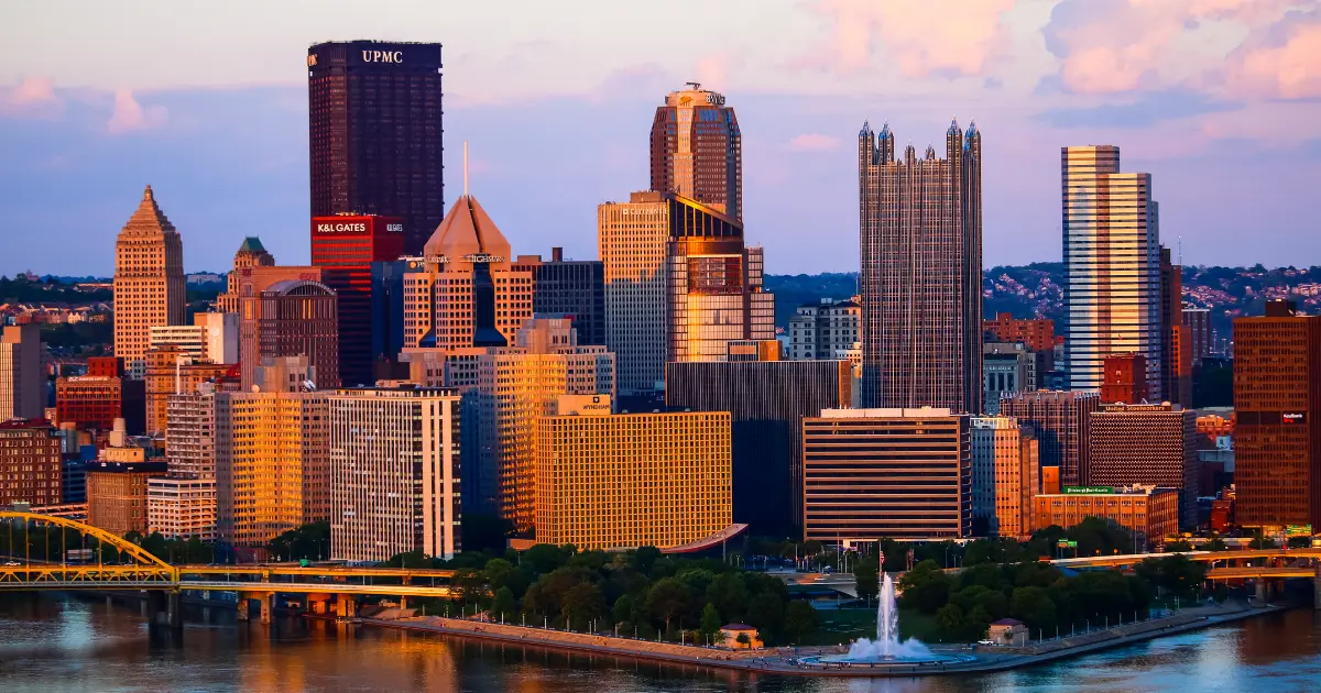 A scenic view of Pittsburgh's skyline at sunset, featuring tall buildings with a mix of modern and classic architecture. The UPMC building is prominent, and the city is bordered by a river with a fountain in the foreground. The sky is partly cloudy with a warm glow from the setting sun.