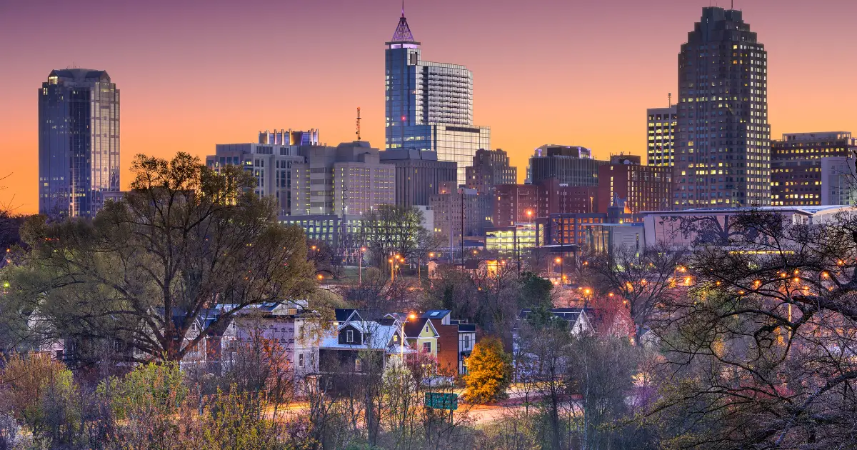 A scenic view of Raleigh, North Carolina's skyline at dusk. The cityscape features tall buildings with lights on, set against a colorful sunset sky. In the foreground, there are trees and residential houses, adding a natural element to the urban scene.
