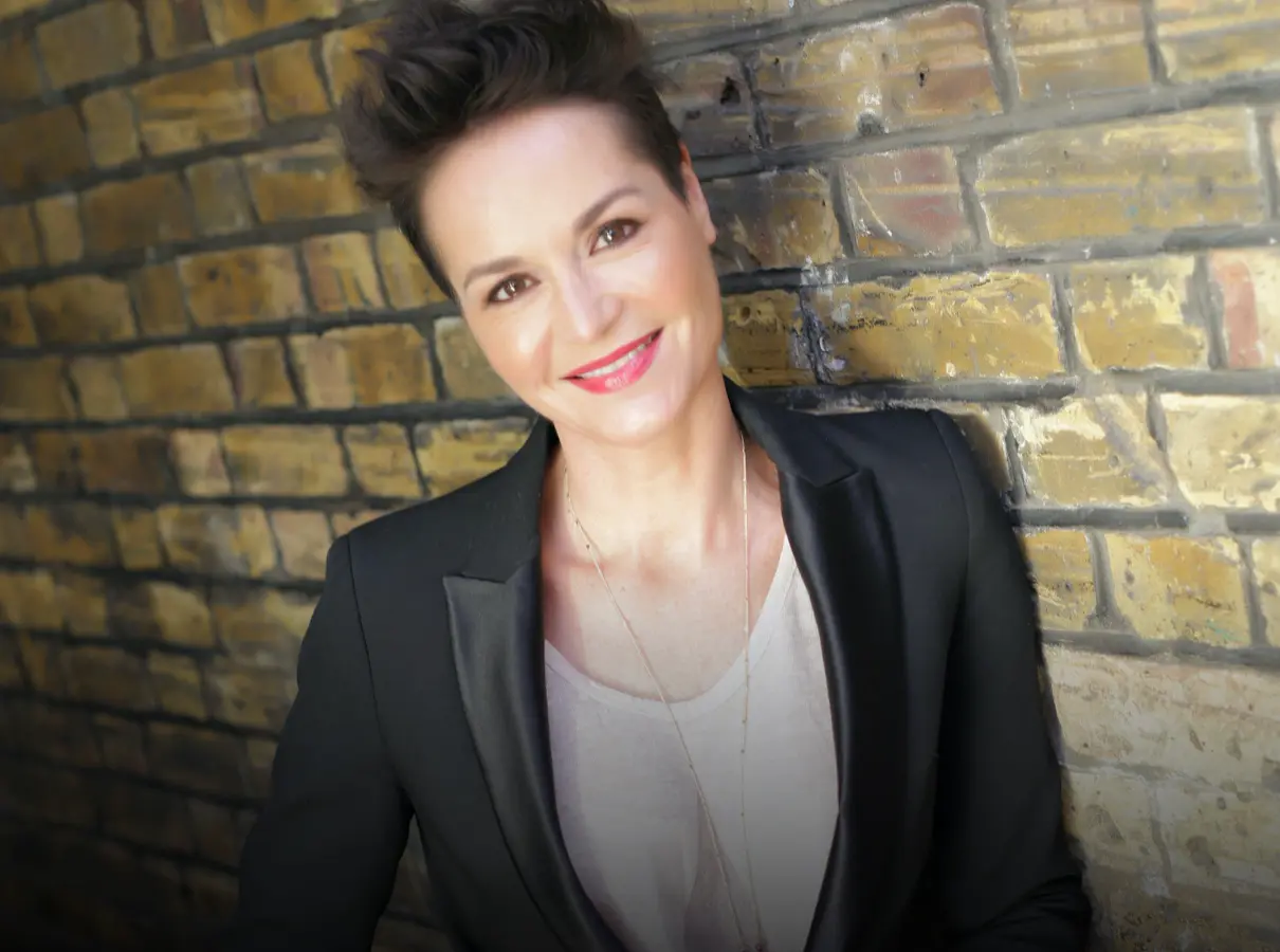 A person with short hair, wearing a black blazer and a light-colored top, smiles while leaning against a brick wall.