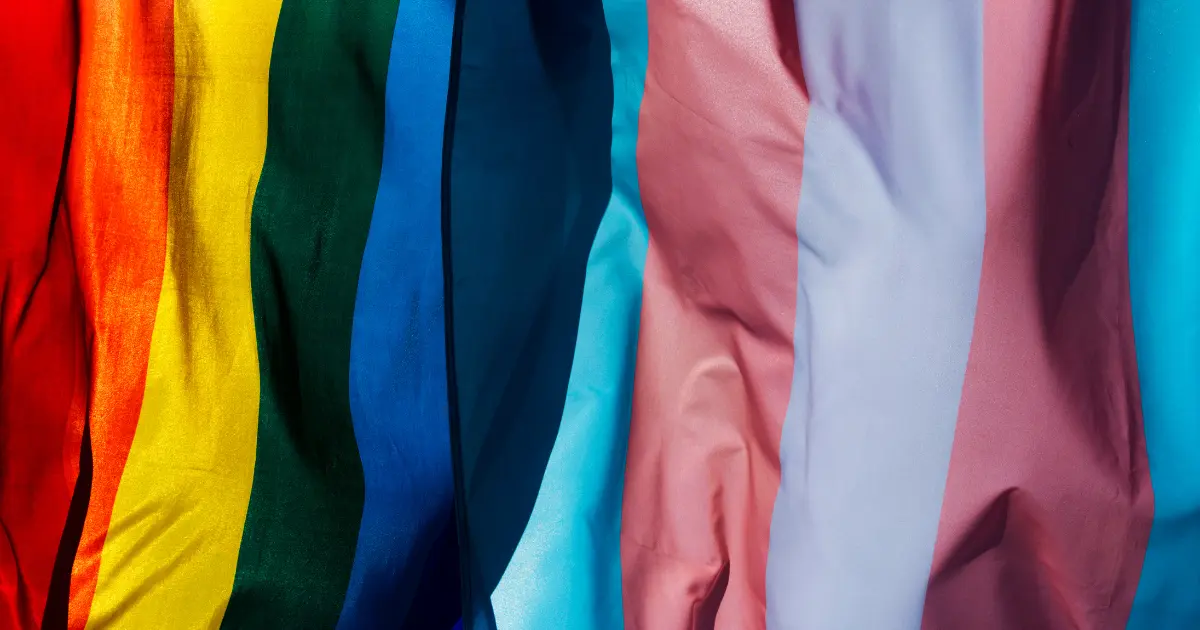 A close-up of colorful fabric strips representing the LGBTQ+ pride flag, including red, orange, yellow, green, blue, and pink hues, draped vertically.