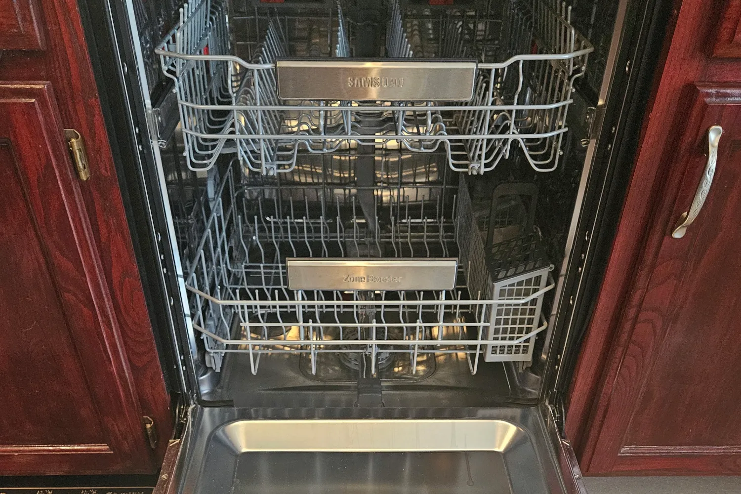 An open, empty Samsung dishwasher built into dark wood kitchen cabinets showing the upper and lower racks