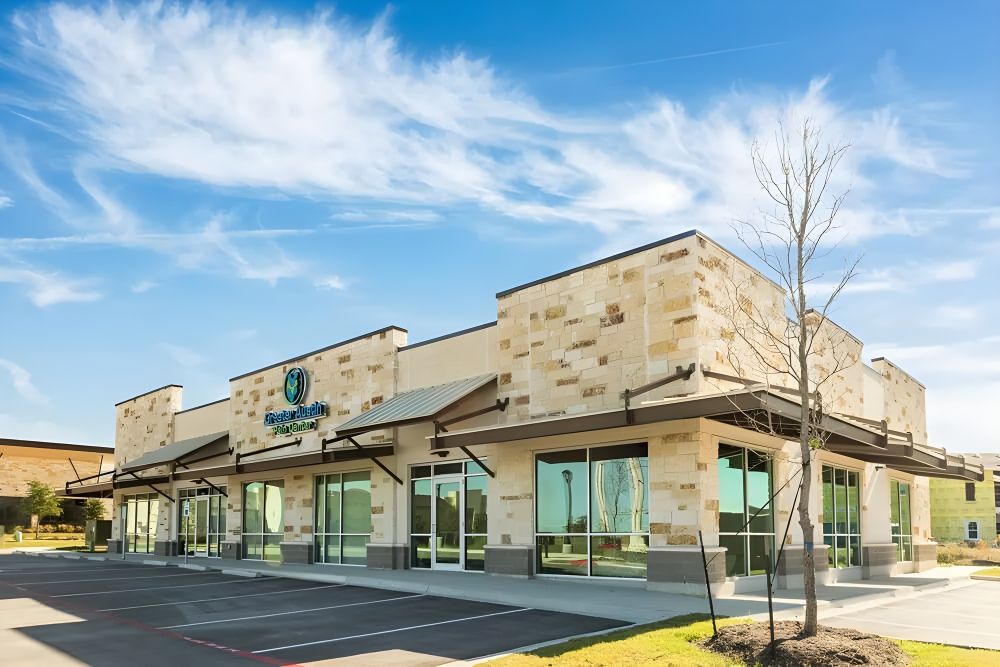 A modern one-story medical office building with stone exterior and large windows, featuring the Greater Austin Pain Center logo on the front, under a bright blue sky with wispy clouds.