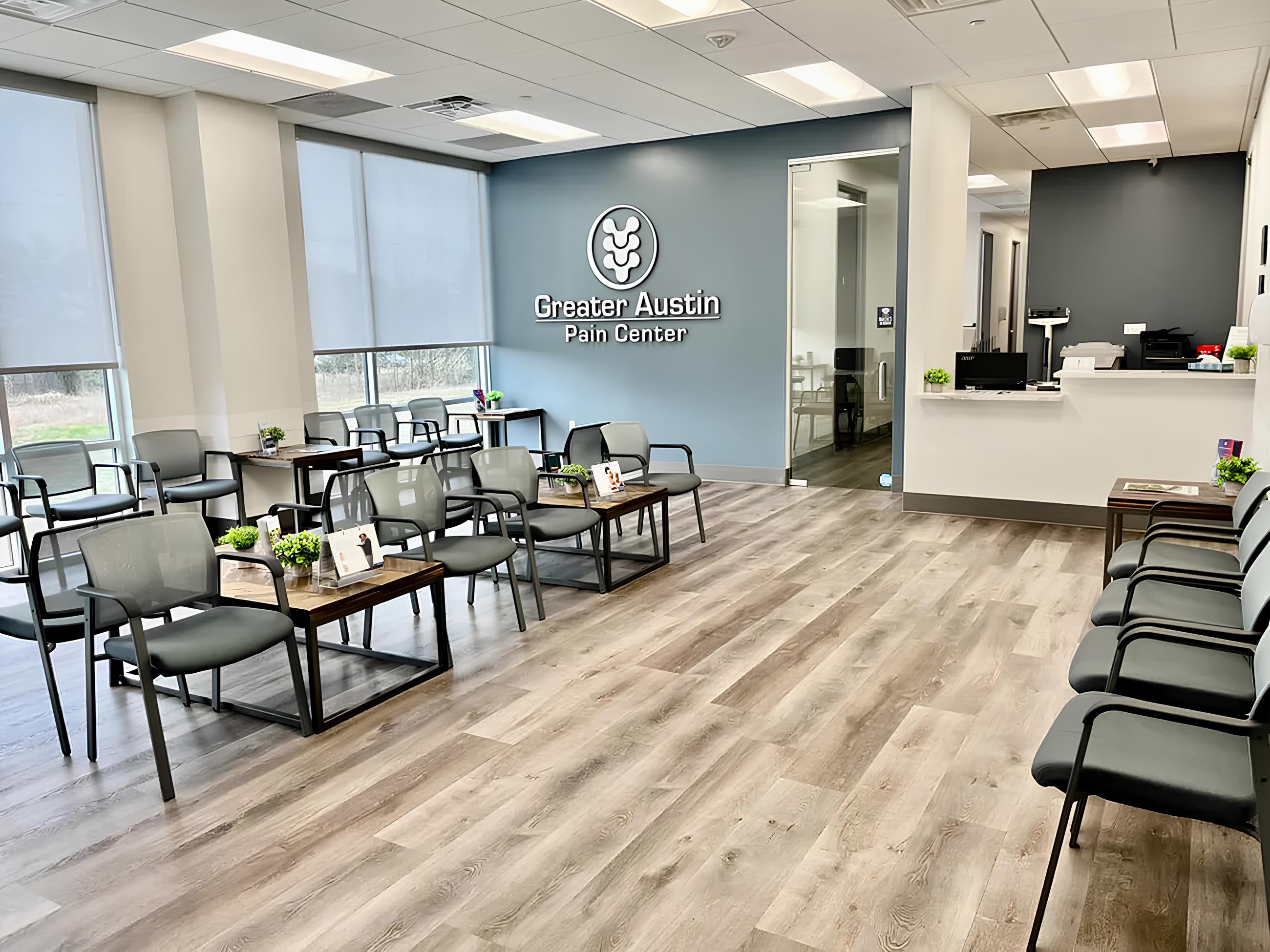 A modern medical office waiting room with gray chairs, small tables with plants and brochures, wood-style flooring, and a reception desk. The wall displays the sign "Greater Austin Pain Center."