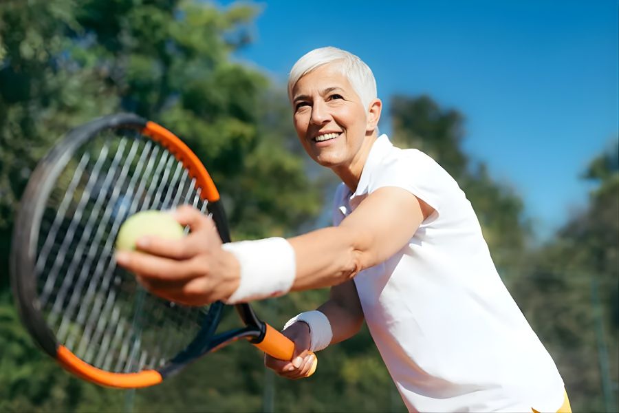 Older woman with short white hair playing tennis, holding a racket and ball.