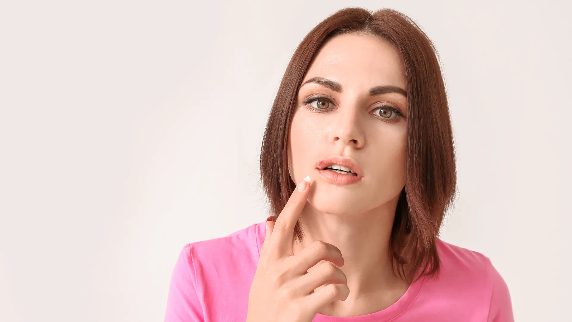 Woman applying cream to chapped lips, wearing a pink shirt, with a neutral background.