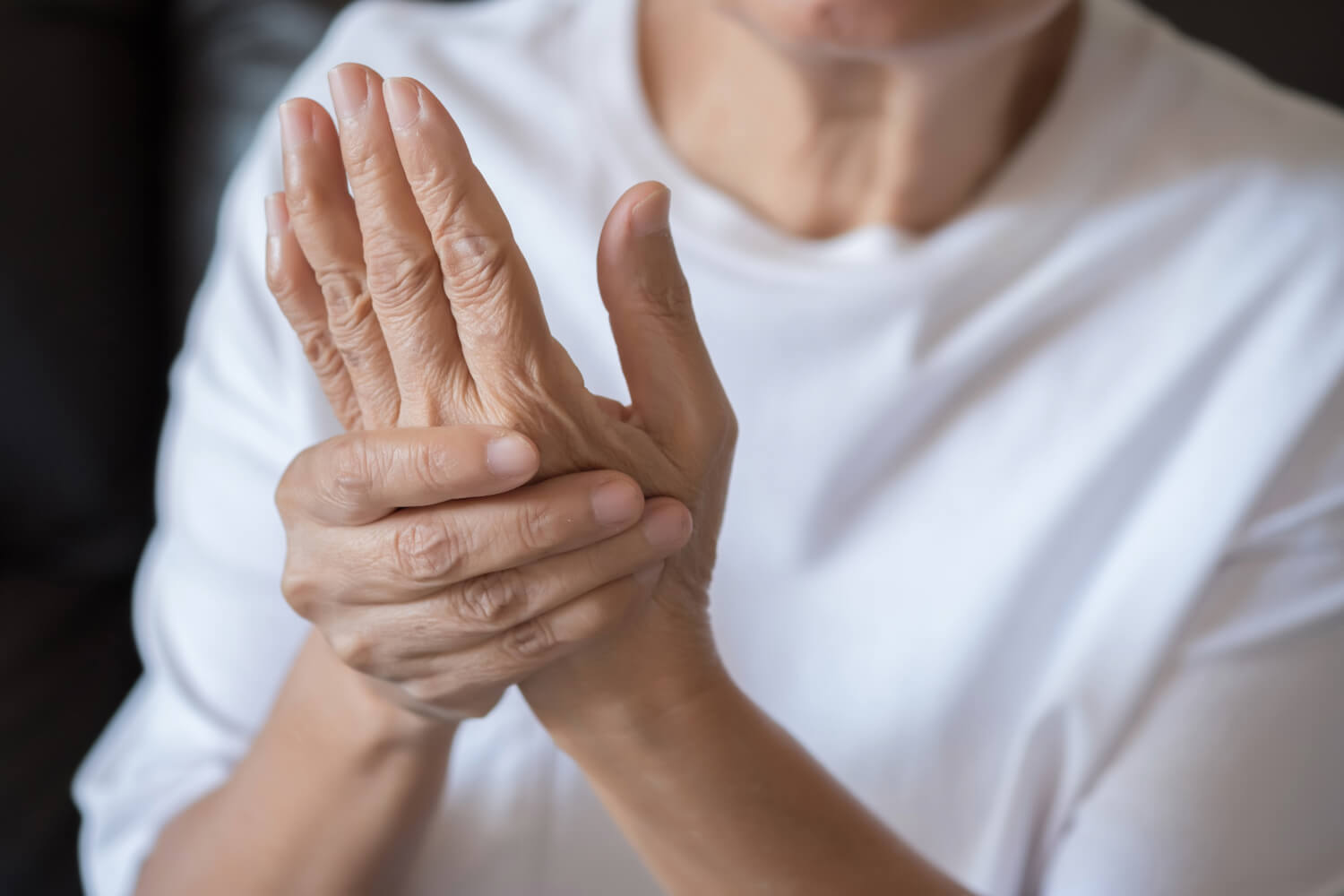 Close-up of an older person wearing a white shirt, holding and massaging their painful hand, possibly indicating discomfort or stiffness from rheumatoid arthritis.