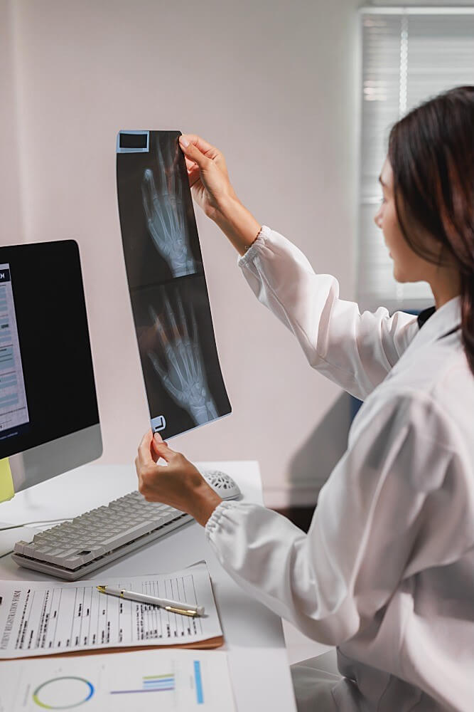 A doctor in a white coat examines hand X-ray films at a desk with a computer and medical documents, likely assessing joint damage or inflammation related to rheumatoid arthritis.