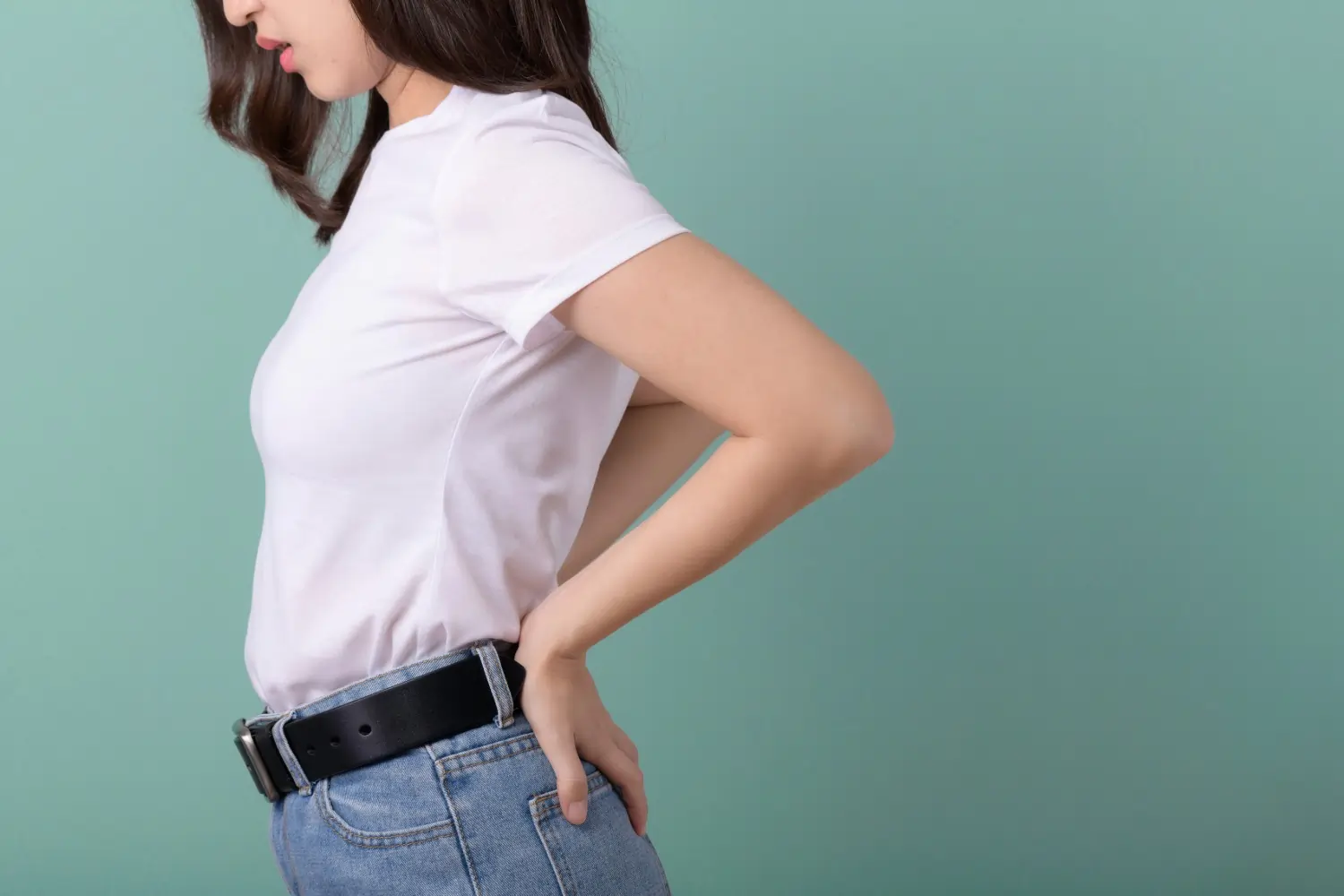 Woman in white shirt and jeans holding her lower back in discomfort against a teal background.