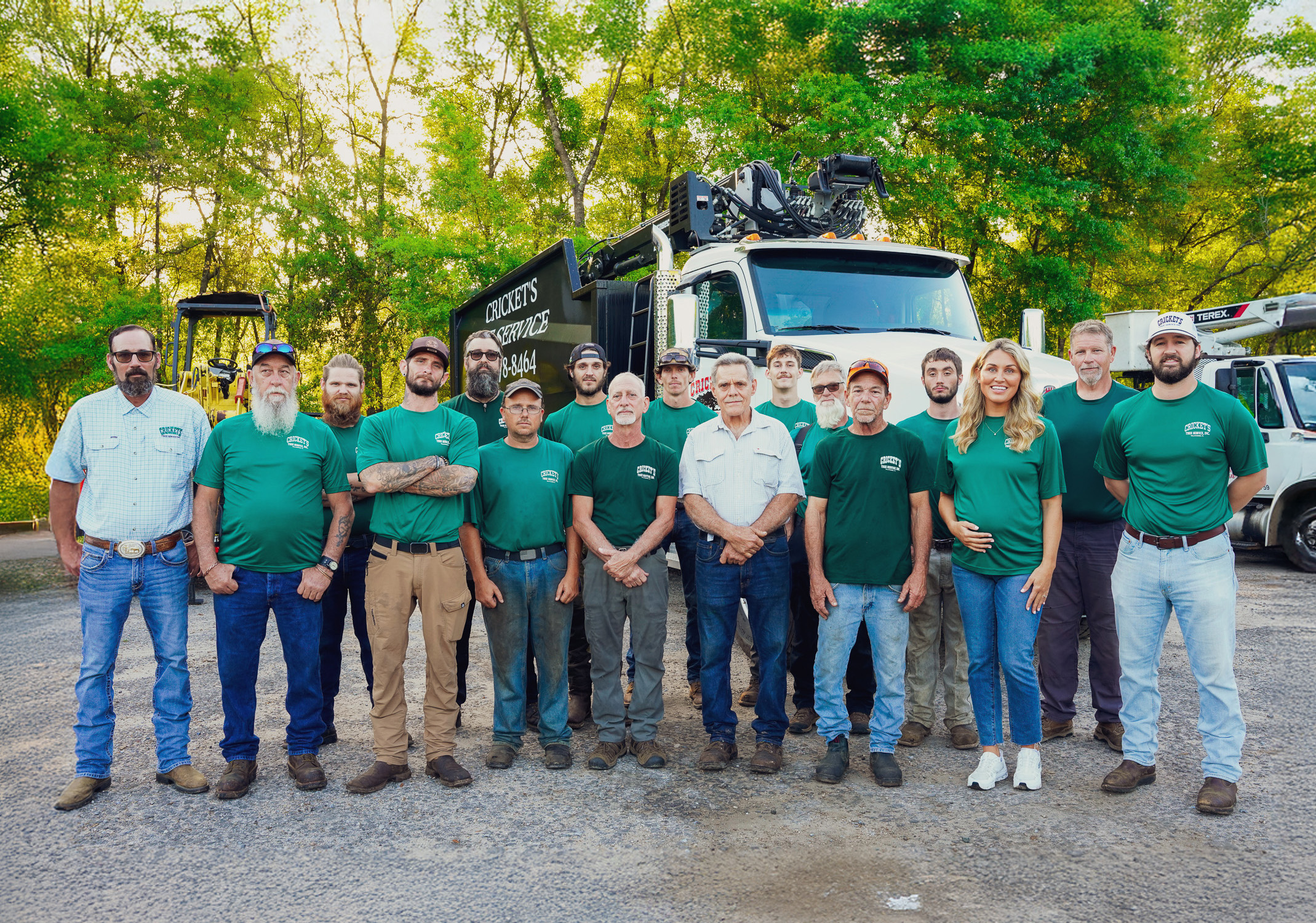 Group photo of fifteen workers standing in front of two large service trucks and construction equipment, surrounded by green trees, with most wearing green shirts with 'Cricket's Service' logo, and one woman in the group.