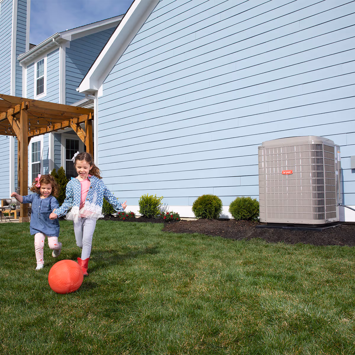 Two children playing with a ball outside with a Bryant HVAC to the side of the frame.