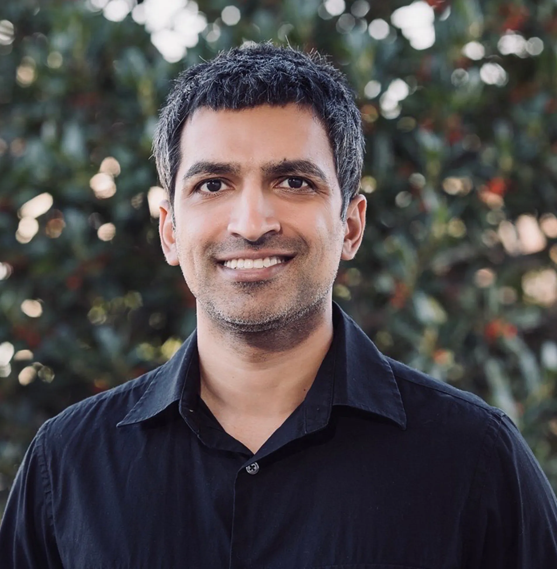 Smiling man with short dark hair and a black collared shirt standing outdoors with blurred greenery in the background.