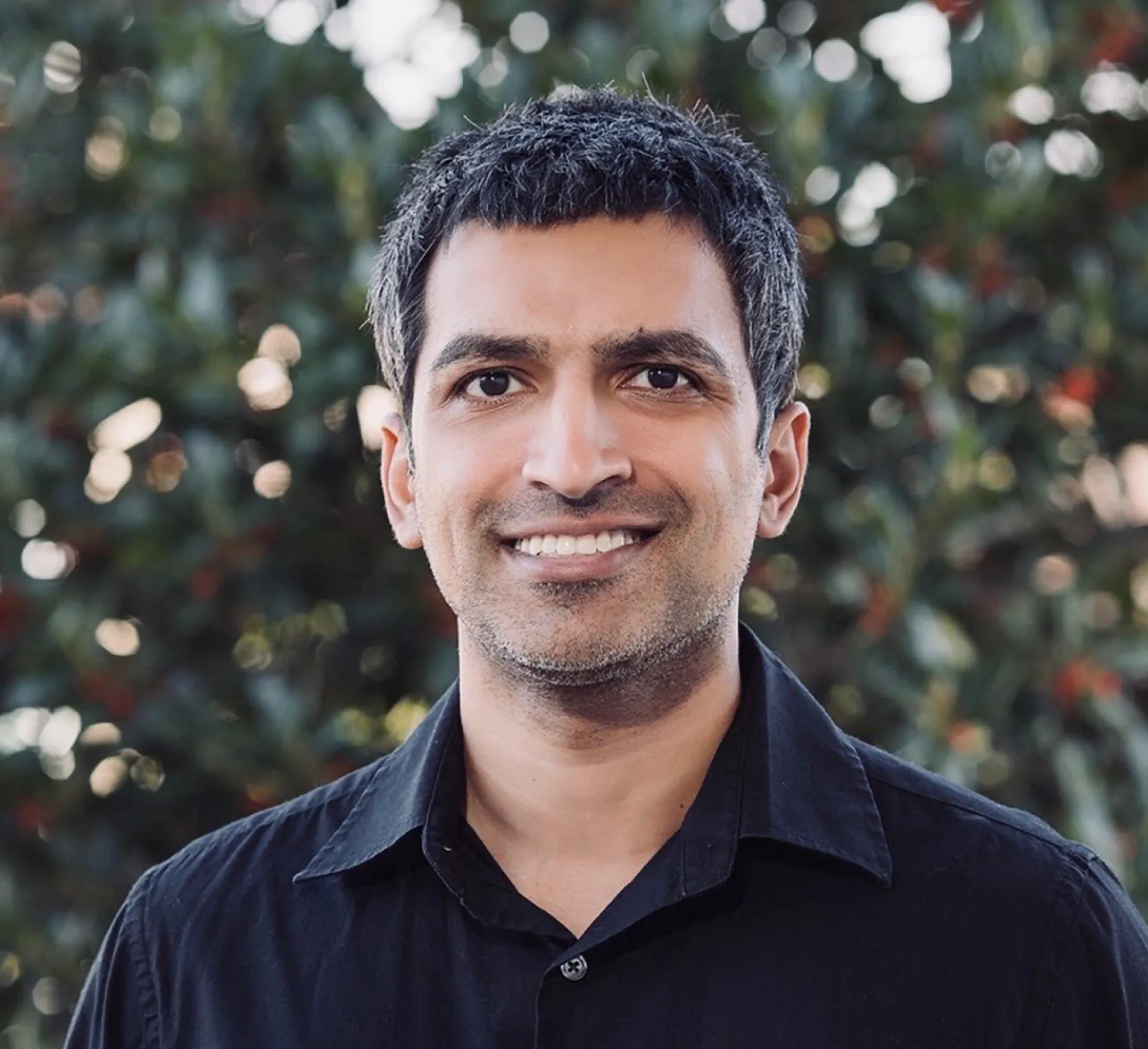 Smiling man with short black and gray hair wearing a black collared shirt, standing outdoors with blurred foliage background.
