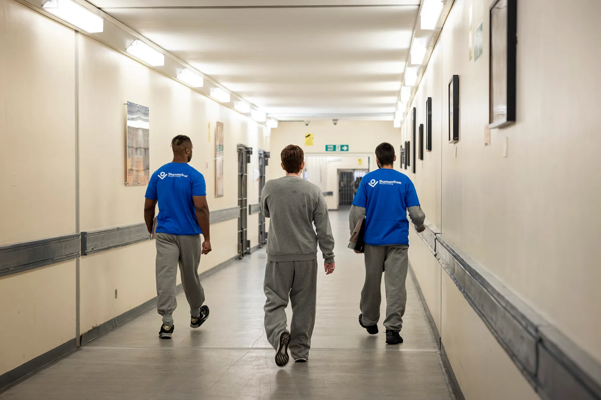 Three people walking away from the camera down a prison corridor, two of them wearing blue Shannon Trust mentor t-shirts.