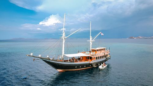 Large traditional wooden sailboat anchored on calm blue sea with people on board and distant islands under a partly cloudy sky.