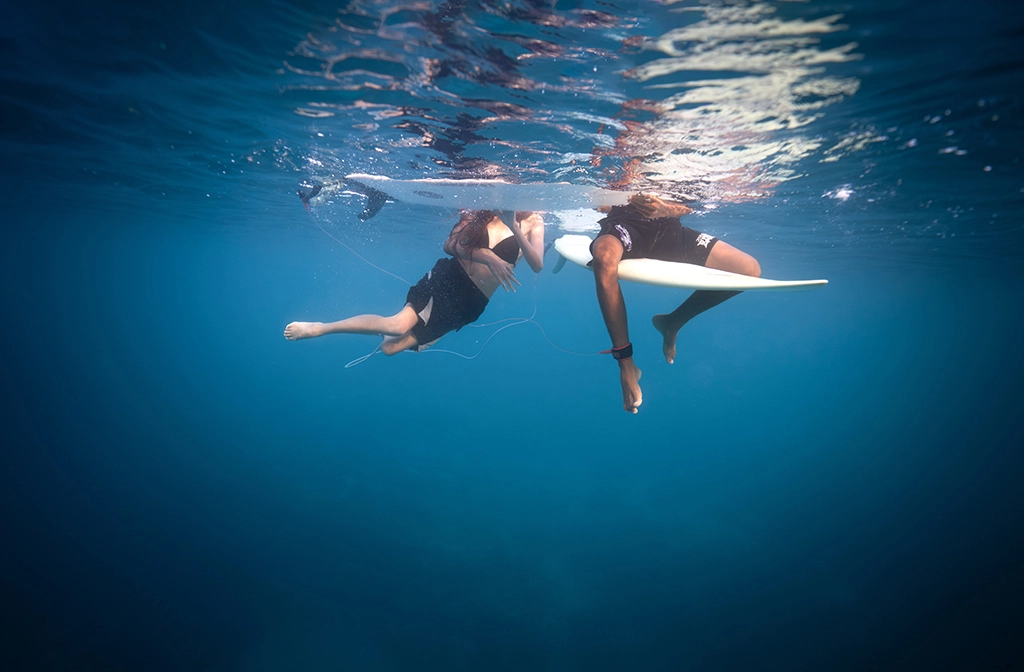 Underwater view of two people with surfboards floating at water surface in clear blue ocean.
