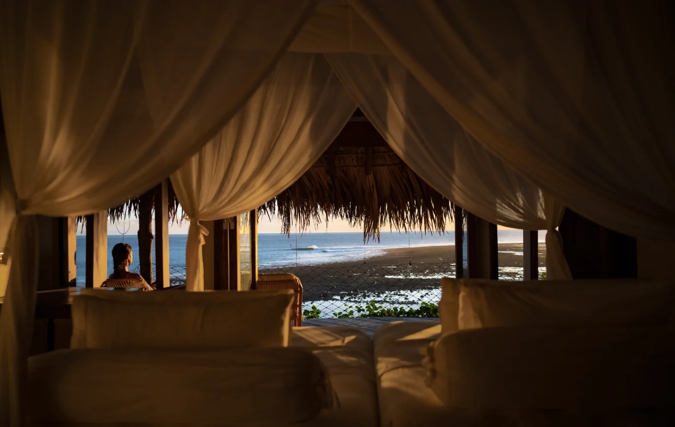 View from a cozy bed with curtains looking out at a person and beach at sunset.