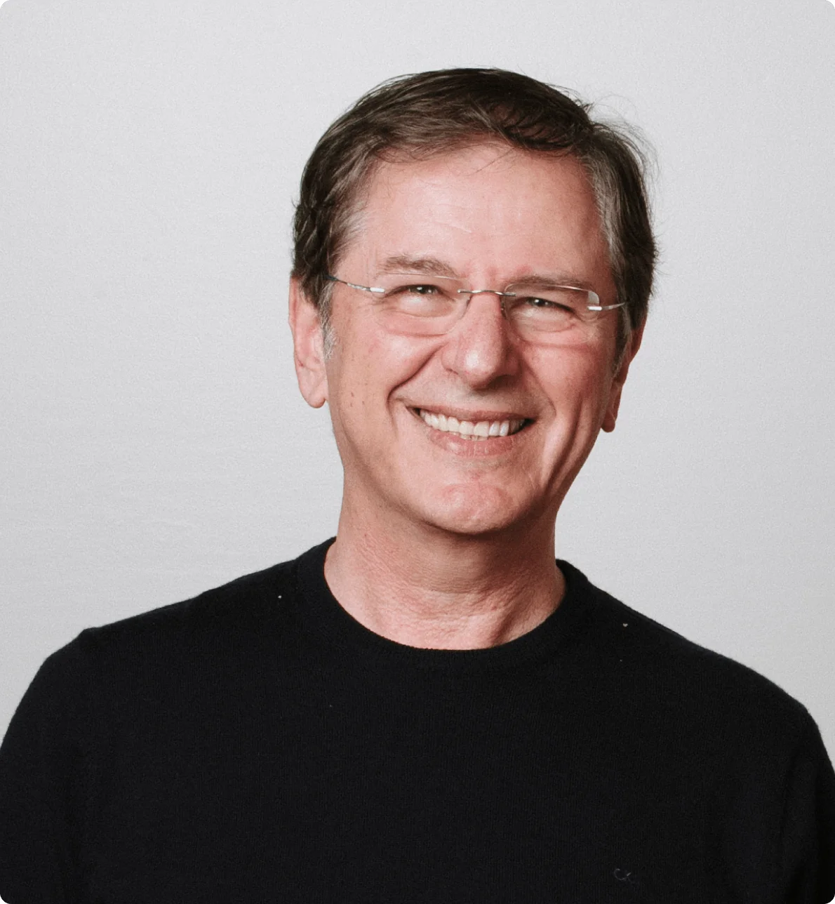 Smiling middle-aged man with short brown hair and glasses wearing a black sweater against a plain light gray background.