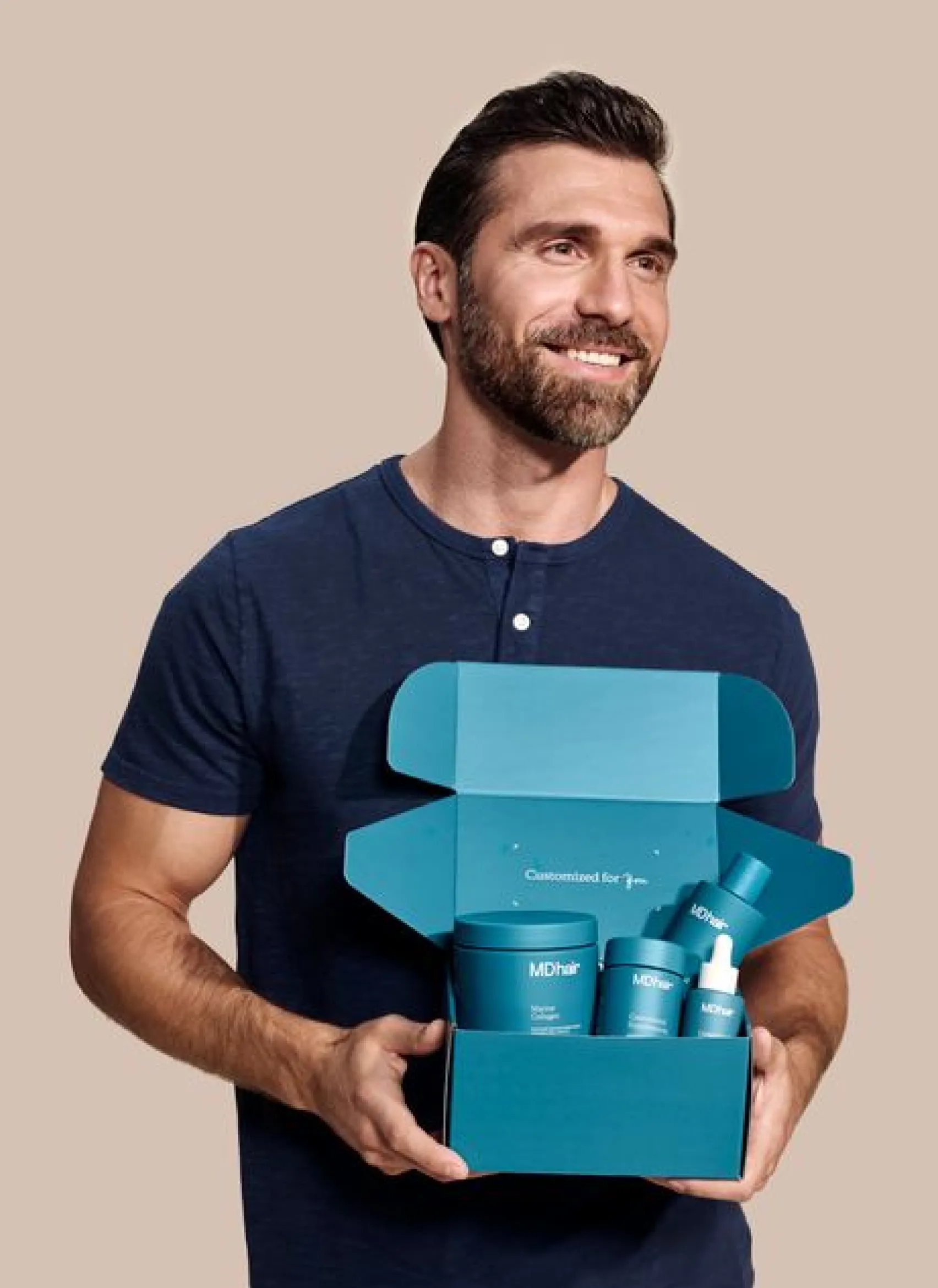 Smiling man with dark hair and beard wearing a navy blue shirt holding an open teal box containing MDhair hair care products including jars, a dropper bottle, and a spray bottle.