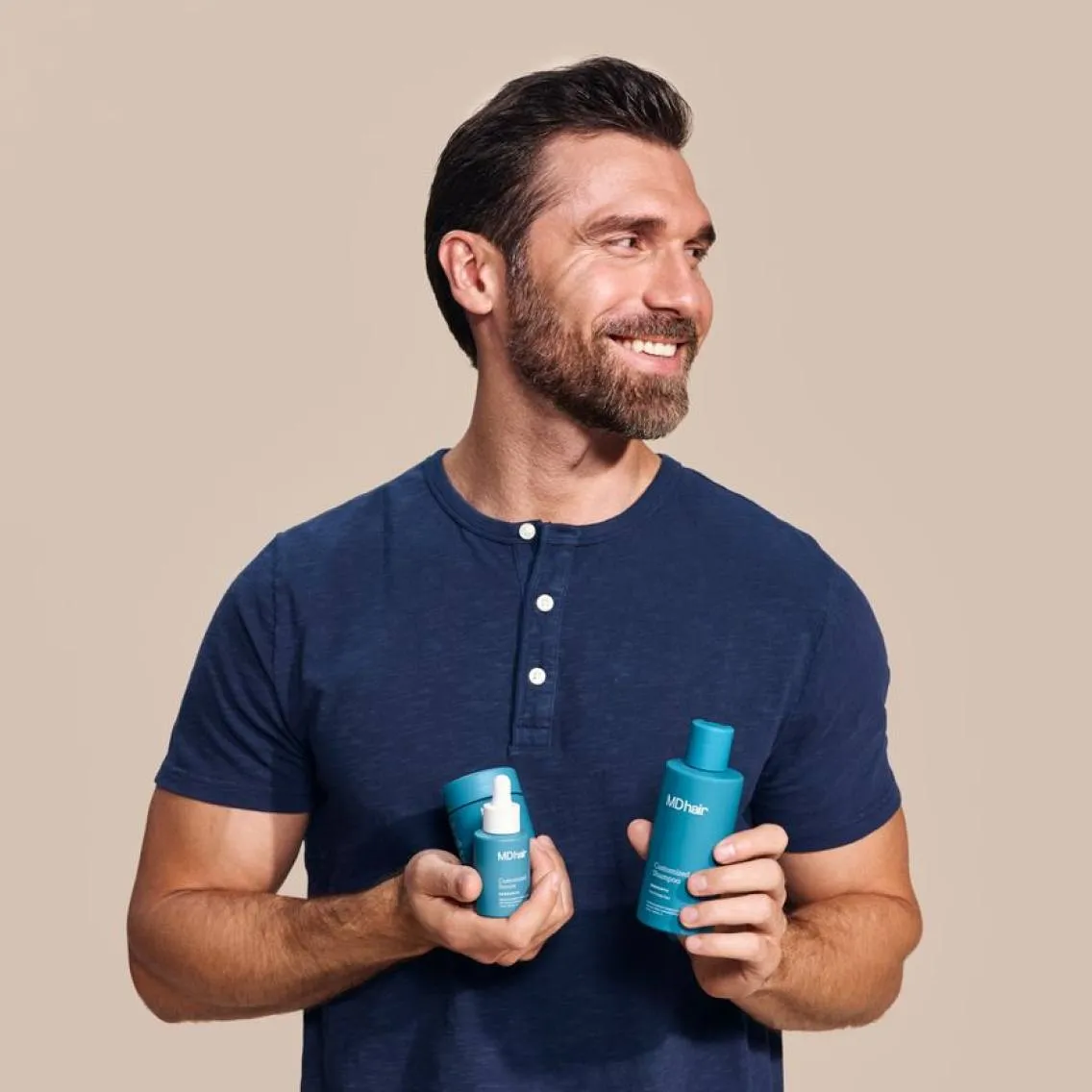 Smiling man with dark hair and beard wearing a navy blue shirt holding three teal MDhair hair care products against a beige background.