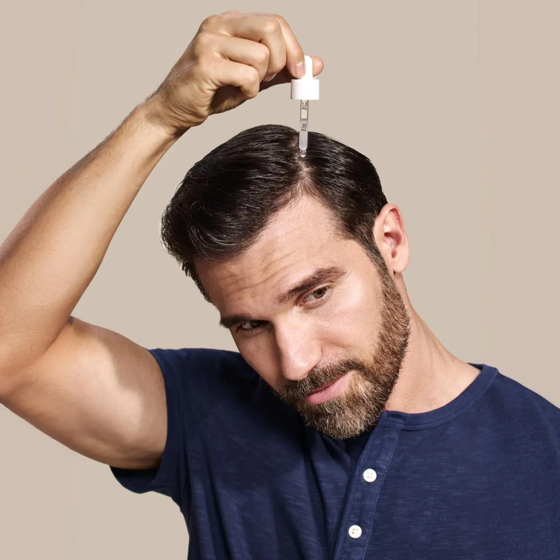 Man with dark hair and beard wearing a navy blue shirt applies scalp treatment using a dropper above the center of his head against a beige background.