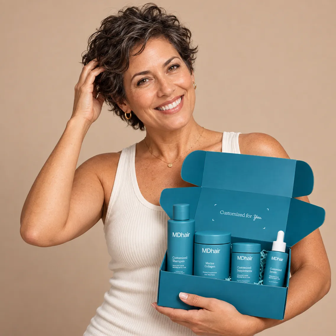 Smiling woman with short curly hair wearing a white tank top holding an open blue box containing MDhair products including shampoo, marine collagen, supplements, and serum, against a beige background.