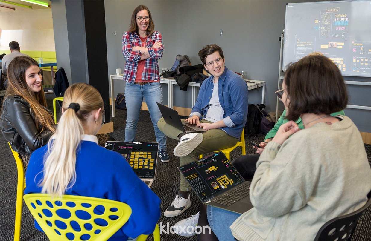 A trainer leads a language course in a group of 5 people in a relaxed atmosphere. Everyone has a laptop or smartphone to participate in the course on the Board.