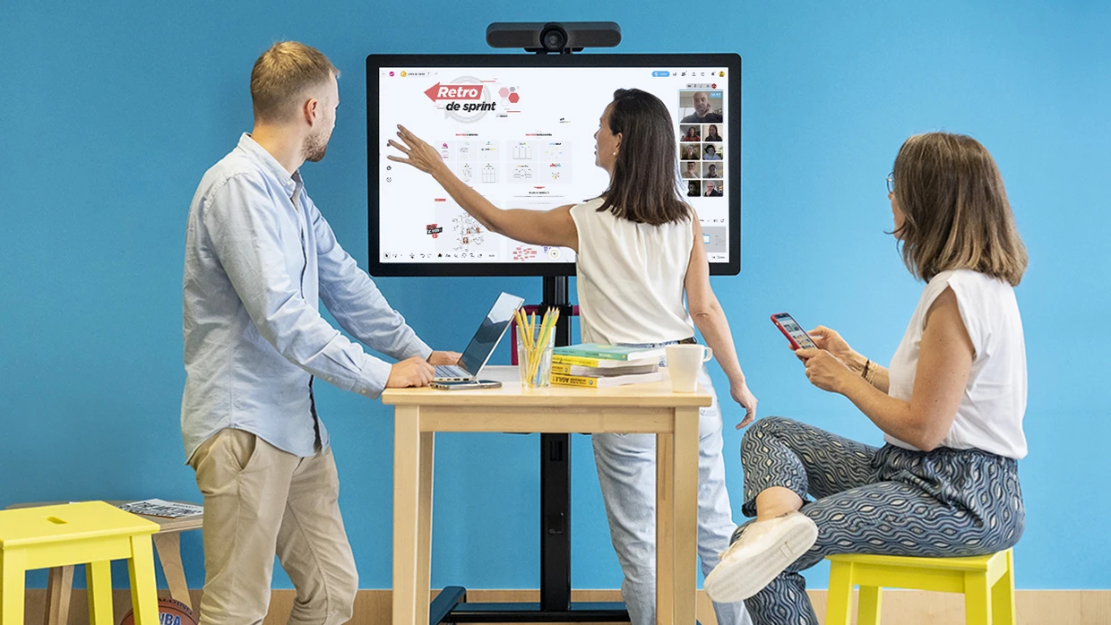 Three people gathered around a large screen displaying a workspace with other people connected via video conference. | Klaxoon