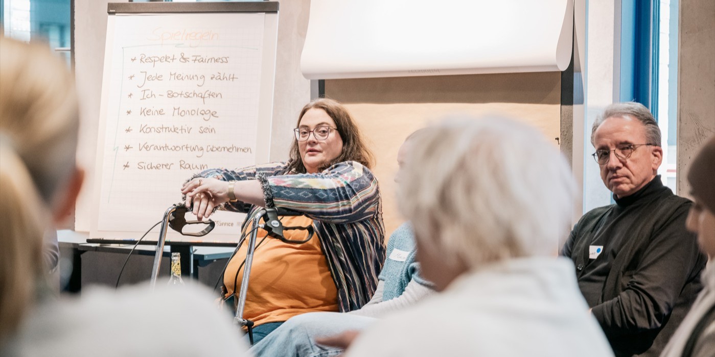 Ein Stuhlkreis mit Menschen und links ein Flipchart. Zwei Menschen - eine Frau mit Rollator und ein Mann - schauen in die Kamera.
