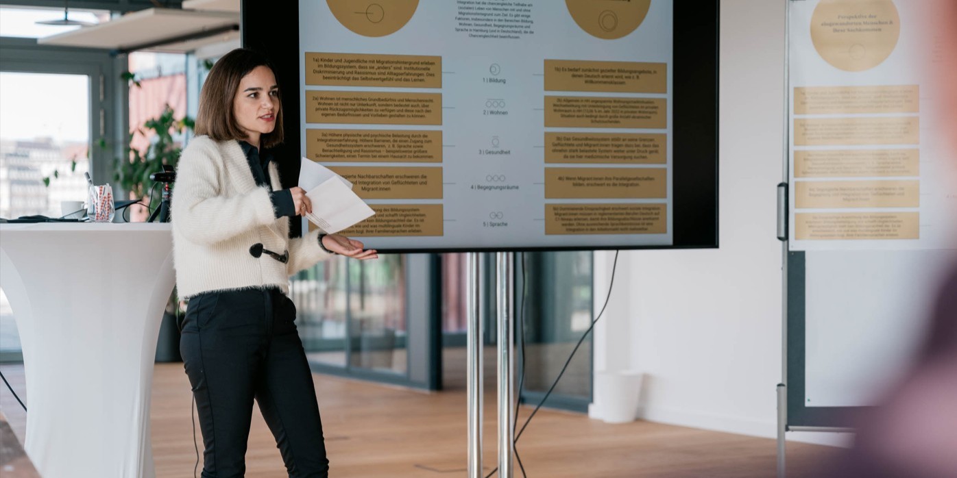 Eine Frau hält eine Präsentation. Sie hat Zettel in der Hand und redet. Rechts neben ihr ist ein Fernseher mit einem Präsentations-Slide.