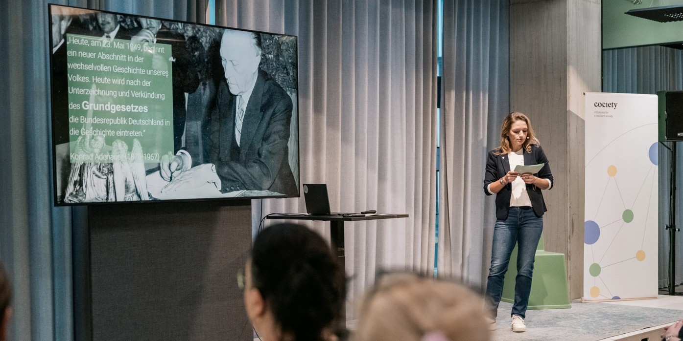 Eine Frau steht auf einer Bühne. Neben ihr ist ein Fernseher mit einem Präsentation-Slide.