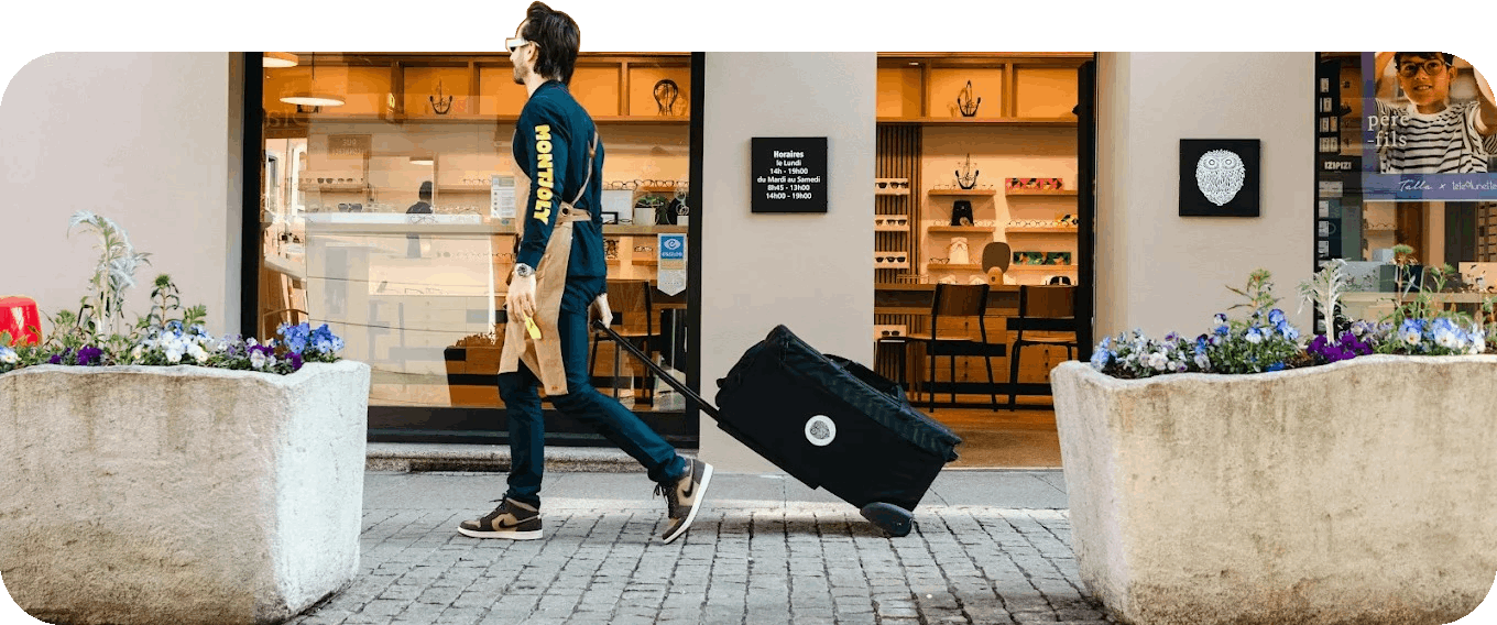 Man wearing a Montpilly sweatshirt and apron walking on cobblestone pavement pulling a black wheeled bag past a store with large planters and flower arrangements outside.
