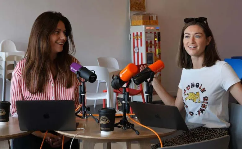 Two women sitting at a round table with laptops and microphones, engaged in a conversation, with coffee cups and a popcorn machine in the background.