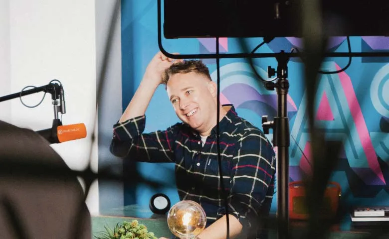 Smiling man in a plaid shirt scratching his head in front of a colorful wall with a microphone to his left and studio lighting equipment visible.